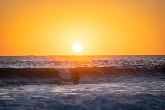 View of a surfer riding a wave during a vibrant orange sunset over the Atlantic Ocean in El Cotillo, Canarias, Spain.