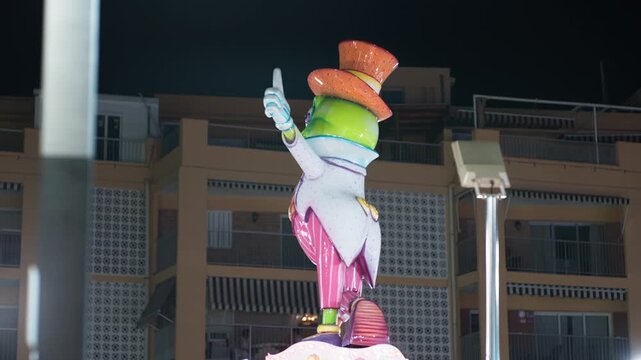 Colorful papier mache festival figure of whimsical cartoon character in top hat, mounted on rotating monument at night, residential buildings behind, fiesta
