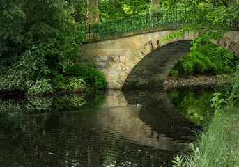 Fototapeta premium The stone Augustenbrucke spanning a tranquil canal within Georgengarten Park in Hannover, Lower Saxony, Germany, surrounded by green foliage.