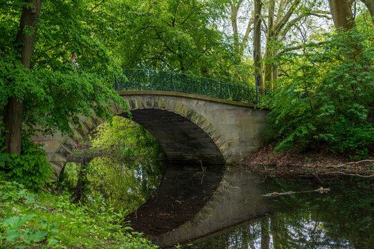 The historic Augustenbrucke reflected in calm water, surrounded by lush green foliage within Georgengarten, Hannover, Lower Saxony, Germany.