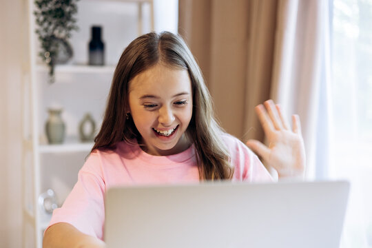 Young girl smiling and waving to laptop screen, enjoying remote communication and online connection from home