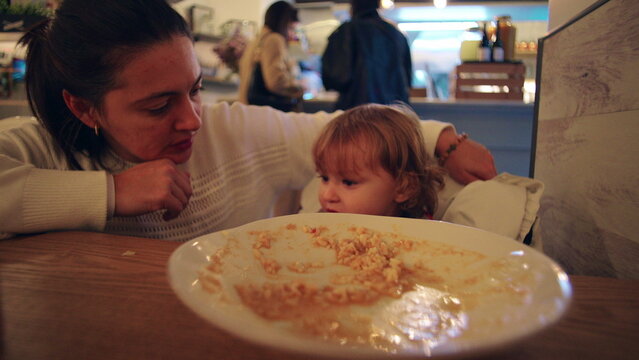 Mother feeding toddler with spoon at restaurant table during family outing sharing meal bonding moment expressing care patience nurturing parenting everyday life scene