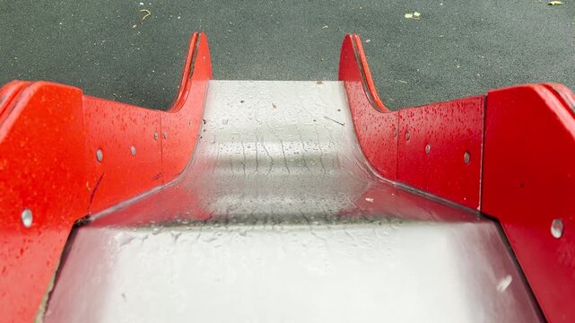 rain puddles sit on a metal slide. A child sliding down creates a loud splash. The wet, empty park brings a lonely and peaceful feeling.