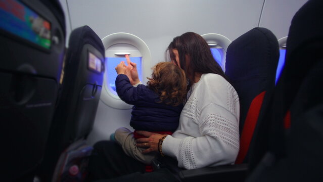 Toddler seated on mother lap opening and closing airplane window shade while exploring cabin evoking curiosity learning and calm anticipation before takeoff