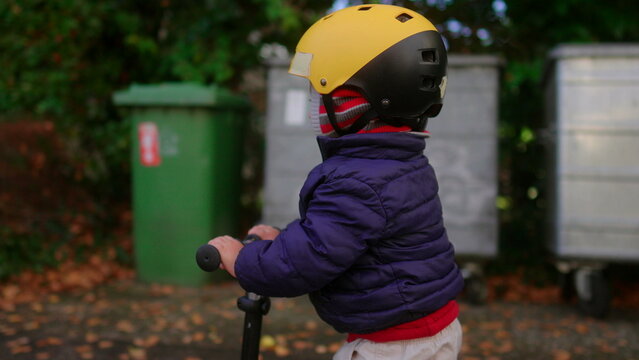 Toddler riding scooter away from camera along autumn sidewalk in residential area wearing helmet practicing balance independence and active outdoor exploration