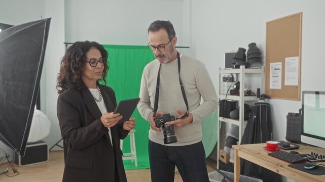 Woman holding tablet while photographer holds camera and reviews images in studio with green screen, softbox and equipment shelving; creative collaboration confidence.