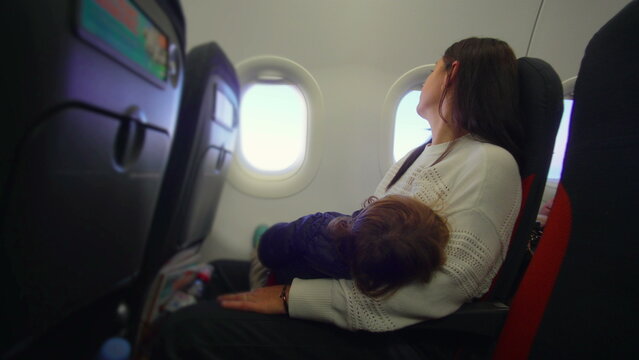 Mother seated on airplane with toddler resting across her lap while waiting for takeoff evoking fatigue tenderness patience and quiet family travel pause