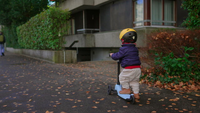 Toddler riding scooter away from camera along autumn sidewalk in residential area wearing helmet practicing balance independence and active outdoor exploration