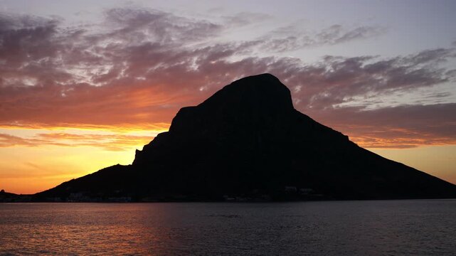 Breathtaking sunset over Telendos, a picturesque island in the Aegean Sea near Kalymnos. Warm golden and orange light bathes the rugged limestone cliffs, tranquil sea, and traditional waterfront
