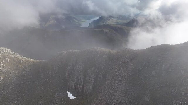 An aerial dolly lateral right shot reveals rugged, mist-shrouded summits in Scotland. The drone glides past craggy terrain, capturing a dramatic, atmospheric vista of the mountainous wilderness.