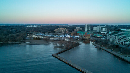 Obraz premium A beautiful high-angle drone photograph over Lake Ontario, capturing the long L-shaped concrete pier at Bronte Harbour, empty winter boat slips, and waterfront buildings under a twilight sky.