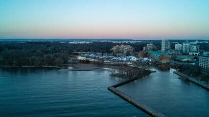 Obraz premium A beautiful high-angle drone photograph over Lake Ontario, capturing the long L-shaped concrete pier at Bronte Harbour, empty winter boat slips, and waterfront buildings under a twilight sky.