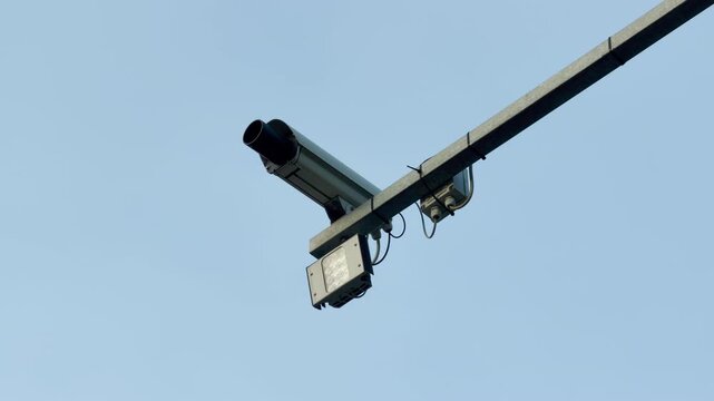 An automated traffic camera is fixed to an overhead metal beam. The device hangs equipped with an infrared flash unit. The system monitors vehicles against a plain, bright sky backdrop.