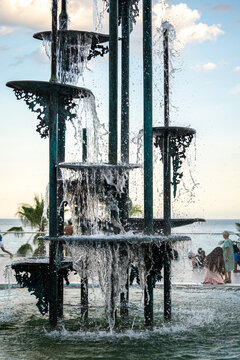 View of a modern tiered bronze fountain with cascading water and people on a promenade by the sea in Monaco, Monaco.