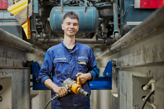 A smiling mechanic in blue overalls works on a truck's suspension using a pneumatic wrench. The workshop is filled with tools, capturing a professional and friendly atmosphere.