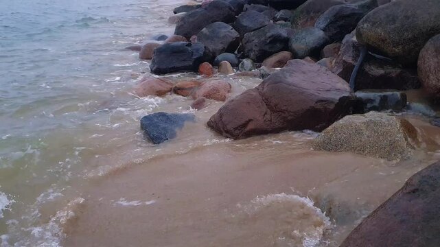Close-up of calm coastal surf water waves breaking on rocky shore with sand beach and huge boulders on Baltic sea coast. Minimalist seaside aquatic pattern natural background.
