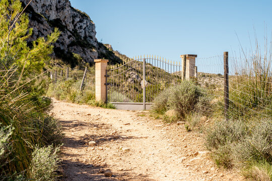View of a metal gate with stone pillars on a dirt path leading into a rocky hillside with green vegetation in Andratx, Balearic Islands, Spain.