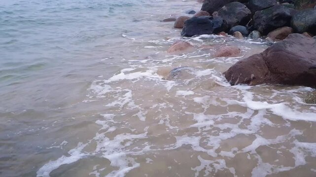 Close-up of calm coastal surf water waves breaking on rocky shore with sand beach and huge stones on Baltic sea coast. Minimalist seaside aquatic pattern natural background.
