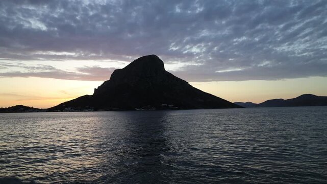 Breathtaking sunset over Telendos, a picturesque island in the Aegean Sea near Kalymnos. Warm golden and orange light bathes the rugged limestone cliffs, tranquil sea, and traditional waterfront