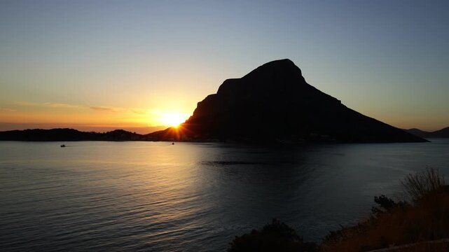 Breathtaking sunset over Telendos, a picturesque island in the Aegean Sea near Kalymnos. Warm golden and orange light bathes the rugged limestone cliffs, tranquil sea, and traditional waterfront