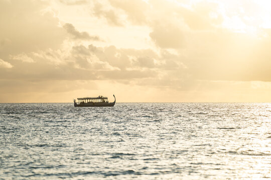 View of a traditional dhoni boat sailing on the calm ocean under a golden sunset sky with soft clouds in Dhigurah, Alif Dhaal Atoll, Maldives.