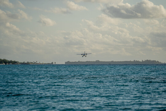 View of a seaplane flying low over the turquoise ocean water near a tropical island with palm trees under a cloudy sky in Dhigurah, Alif Dhaal Atoll, Maldives.
