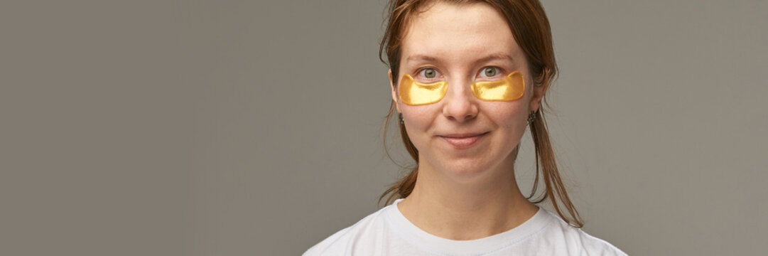 Young Woman Wearing Gold Eye Patches Smiling With Subtle Grin In Wide Horizontal Studio Composition Wearing Casual White T Shirt Against Smooth Gray Background.