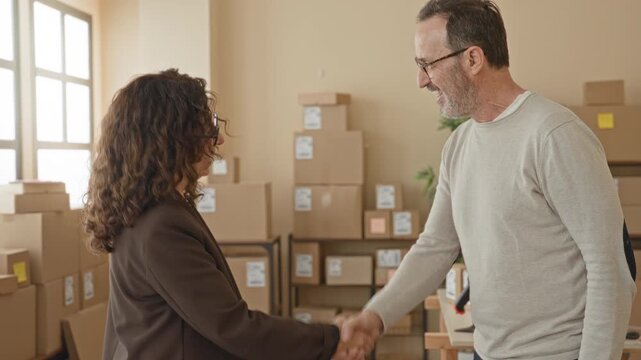 Woman in glasses shakes hand with man amid stacked cardboard boxes and shipping labels in a building storage office; trust partnership.