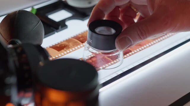Photographer Hand Holding Magnifier Loupe Inspecting Analog Film Negative Lightbox Studio. A professional creator holds a 10x magnification loupe tool over a strip of 35mm celluloid frames