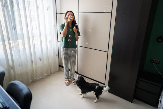 Chinese woman blowing soap bubbles while dog watches indoors