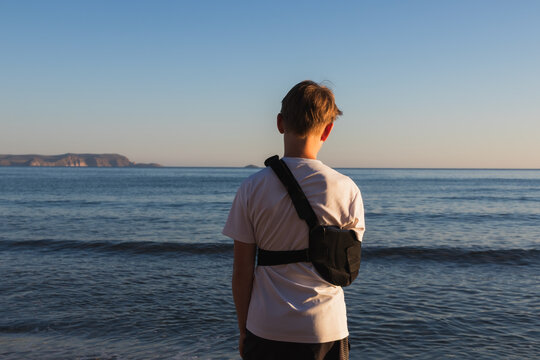 Close up back view of a boy with a broken arm wearing a black medical shoulder sling bandage on a beach looking at the sea