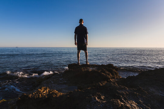 Rear view of a 43 year old man standing on dark rocks by the ocean, enjoying the scenic sea view during summer vacation in Crete, Greece
