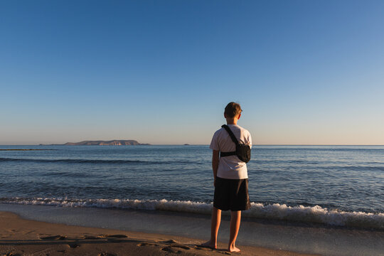 An 11 year old boy with a broken arm in a medical shoulder sling immobilizer bandage standing on a sandy beach looking at the sea during golden hour
