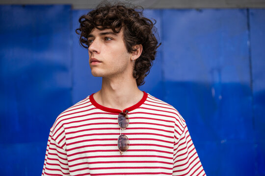 Curly-haired man in striped shirt posing by blue wall