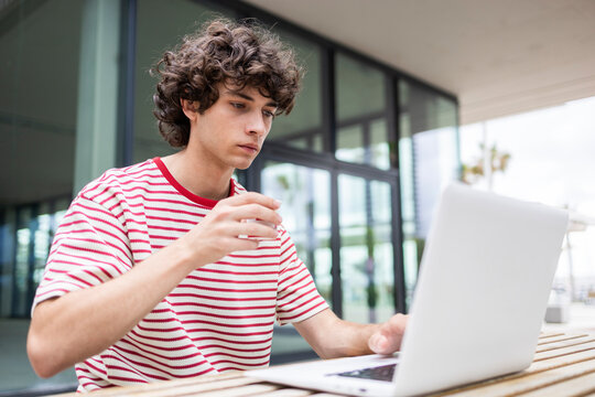 Curly-haired man using laptop outdoors with coffee break