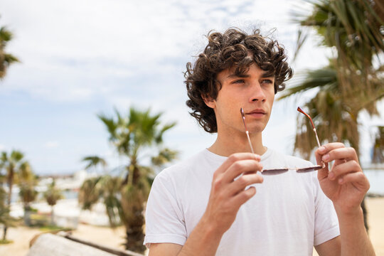 Curly haired man holding sunglasses at a beach promenade