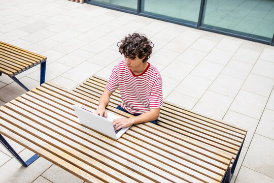 Curly-haired man working on laptop at outdoor bench