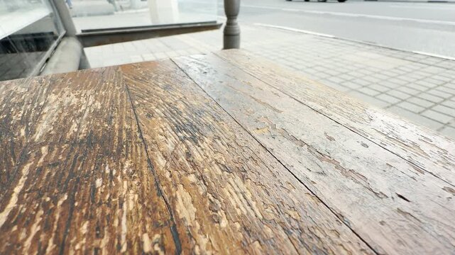 A worn wooden bench surface shows peeling paint and a rough texture. In the blurry background, a large white bus stands on the paved urban road. The public transit stop echoes with roar of traffic.