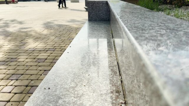 A smooth, reflective marble stone ledge sits beside an urban walkway. In the blurry distance, pedestrians stroll across the sunlit cobblestone square.