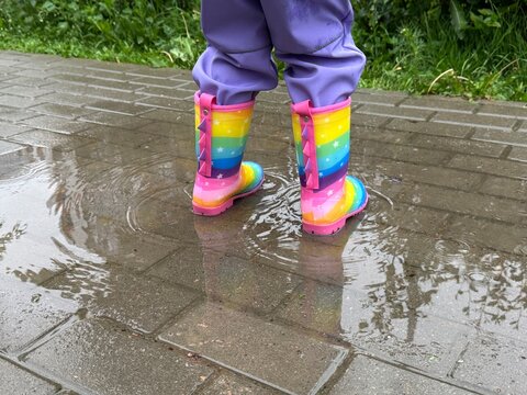 Children's feet stomp in a puddle, close-up, toddler girl walks in multicolor rainbow boots in a puddle in the rain, summer, autumn, spring.
