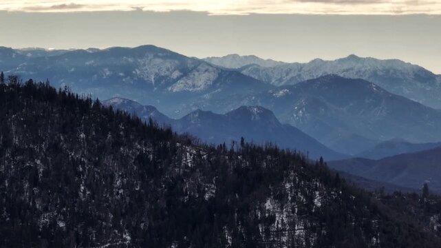 Aerial view of forested mountains with patches of winter snow under a cloudy sky in California