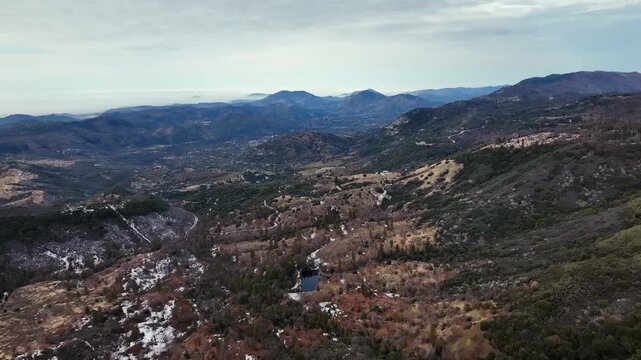 Aerial view of forested mountains with patches of winter snow under a cloudy sky in California