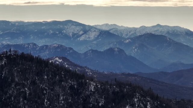 Aerial view of forested mountains with patches of winter snow under a cloudy sky in California