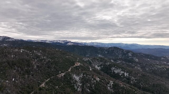 Aerial view of forested mountains with patches of winter snow under a cloudy sky in California