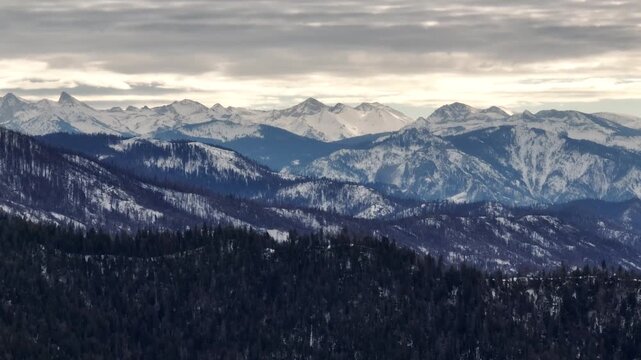 Aerial view of forested mountains with patches of winter snow under a cloudy sky in California