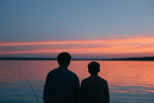 Father and son silhouette fishing together at sunset by the lake, calm water reflecting vibrant colors of the sky, creating a serene outdoor scene