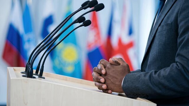 Close up footage of a diplomat standing at a wooden podium with microphones, clasping hands during an international political conference with global flags in the background.