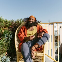 Obraz premium Boho styled man with headband relaxing on a sunny balcony chair holding coffee for bohemian lifestyle background and social media wellness design