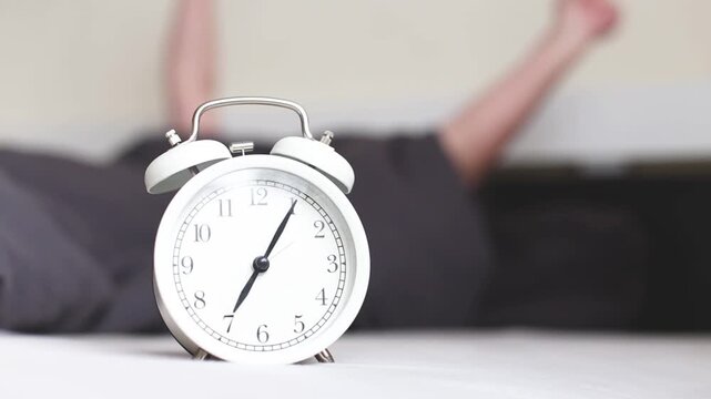 Faceless female person waking up early in the morning lying in her bed under cozy blanket in modern bedroom with alarm clock and stretching arms. Good morning, awake and new day concept.