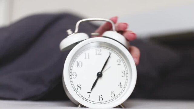 Sleeping person lying on the bed in bedroom and stopping alarm clock on bedside table at home. Wake up in morning of working day. Young child reaching for alarm clock to switch it off